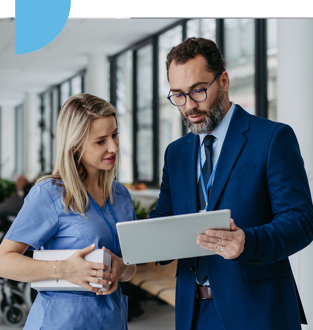 Professional Consultation in Medical Setting A female healthcare professional in scrubs and a male business professional in a blue suit are reviewing information on a digital tablet. They are standing in a modern, bright clinical or hospital hallway, engaged in a collaborative discussion.